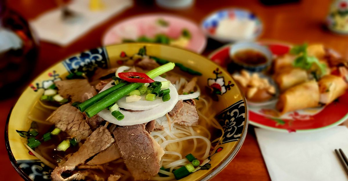 Eine Schüssel vietnamesische Pho-Suppe mit frischen Kräutern neben japanischen Sushi-Rollen auf einem Tisch