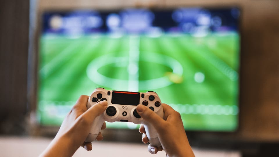 Zwei Hände halten einen Gamecontroller vor einem Fernseher mit laufendem Spiel