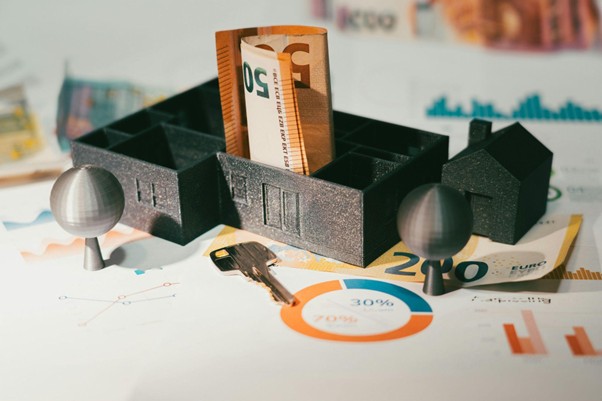 Ein 3D-gedrucktes Hausmodell mit herausstehenden 50-Euro-Scheinen ist auf Diagrammen und Grafiken platziert und symbolisiert die Anliegen der Frankfurter Eigenheimbesitzer. Daneben befinden sich zwei Schlüssel und kleine runde Objekte, die an Bäume erinnern, während auf Blättern Prozentzahlen und Eurosymbole angezeigt werden.