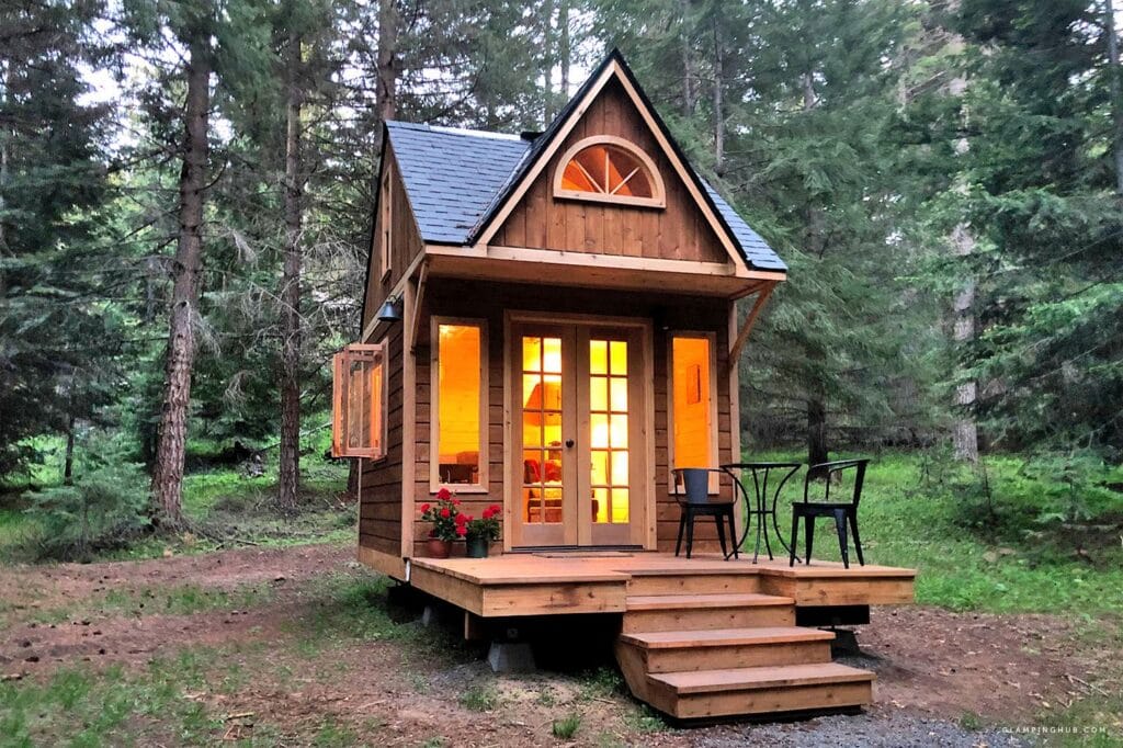 Eine kleine Holzhütte mit warmen Lichtern, großen Fenstern und einer Veranda mit zwei Stühlen und einem Tisch liegt inmitten hoher grüner Kiefern - eines der charmanten 24 qm großen Schlafbodenhäuser, die man in Frankfurts Gärten entdecken kann.