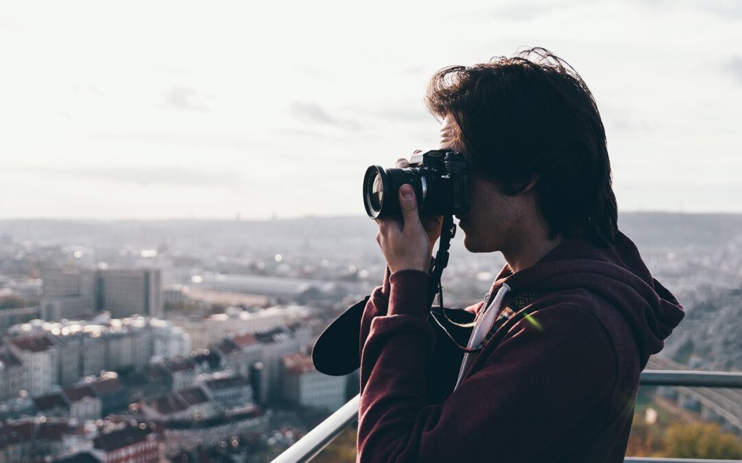 Eine Person mit schulterlangem Haar fotografiert mit einer Kamera von einem hohen Aussichtspunkt aus, von wo aus sie die weitläufige Stadtlandschaft Frankfurts unter einem bewölkten Himmel überblickt. Sie trägt einen kastanienbraunen Kapuzenpullover und hält die Kamera ans Auge. So macht sie Instagram-würdige Fotos.