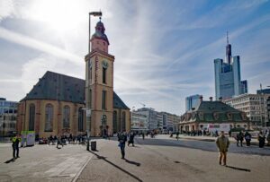 Ein belebter Platz in Frankfurt am Main. Das Bild zeigt die St. Katharinenkirche mit ihrem großen Glockenturm auf der linken Seite, moderne Wolkenkratzer im Hintergrund und zahlreiche Menschen, die unter einem sonnigen Himmel auf dem Platz herumlaufen und sitzen, was das dynamische Leben und die lebendige Gemeinschaft von Wohnen dort veranschaulicht.