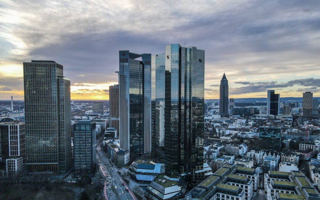 Luftaufnahme der Frankfurter Skyline mit hohen, modernen Wolkenkratzern mit Glasfassaden. Im Hintergrund ist der Sonnenuntergang zu sehen, am Himmel ziehen Wolken auf. Im Vordergrund sind belebte Straßen und kleinere Gebäude zu sehen, darunter eine bepflanzte Dachterrasse in der Nähe der Krypto-Regulierungsbehörde.