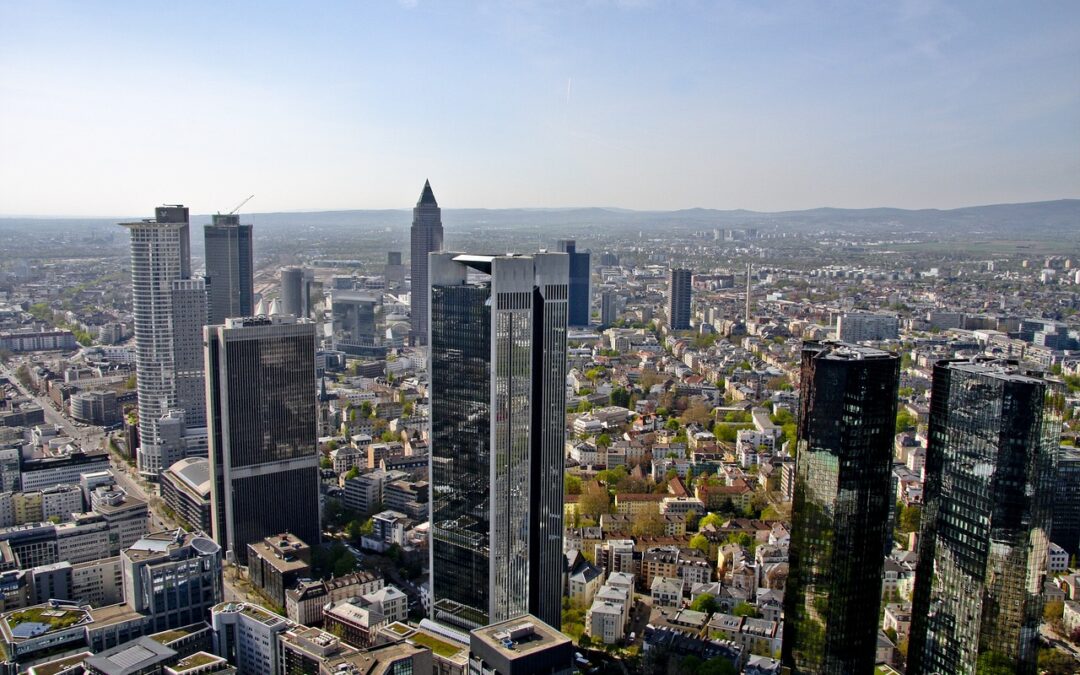 Luftaufnahme einer geschäftigen Skyline in Deutschland, die moderne Wolkenkratzer neben Wohn- und Geschäftsgebäuden zeigt. Die ausgedehnte Landschaft bietet üppige Grünflächen und ferne Hügel unter einem klaren blauen Himmel, ganz wie die lebendigen Welten von Online-Spielen.