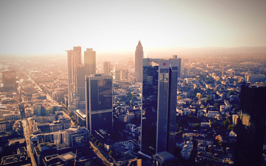 Luftaufnahme einer Skyline bei Sonnenuntergang mit mehreren hohen Wolkenkratzern, darunter ein markanter Turm mit spitzer Spitze. Die Gebäude werden vom goldenen Licht beleuchtet und bilden einen Kontrast zur umgebenden Stadtlandschaft und den fernen Bergen unter einem dunstigen Himmel. Dies fängt Frankfurts kulturelle Highlights ein.
