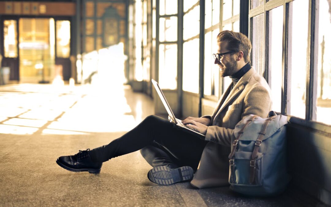 Eine Person auf Geschäftsreise sitzt an großen Fenstern und benutzt einen Laptop. Sie trägt Mantel und Brille und hat einen Rucksack neben sich. Sonnenlicht strömt durch die Fenster und schafft eine warme, helle Atmosphäre – perfekt, um auf Reisen konzentriert zu bleiben.