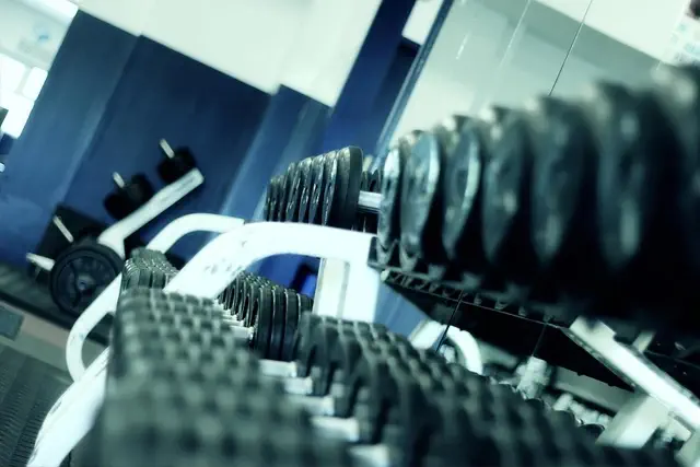 Eine Nahaufnahme einer Reihe schwarzer Hanteln, die auf einem Ständer im Lucius Fit Gym aufgereiht sind. Der Hintergrund ist in Blau und Weiß gehalten und zeigt zusätzliche Fitnessgeräte und Spiegelungen im Spiegel. Das Bild betont eine saubere, organisierte Fitnessstudioumgebung.