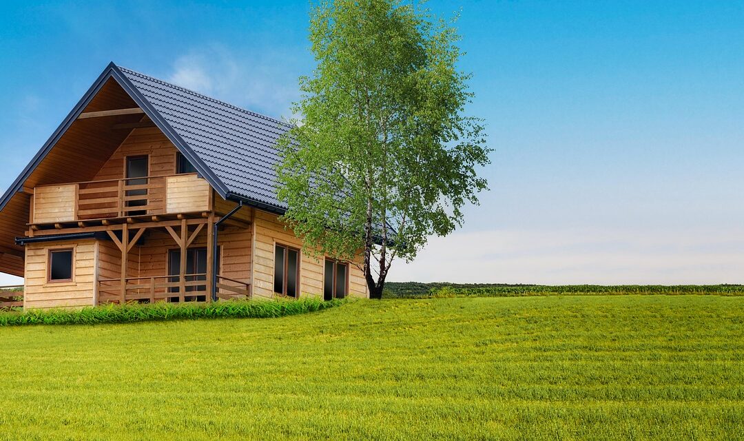 Ein gemütliches Ferienhaus aus Holz mit steilem blauem Dach steht auf einer Wiese unter dem klaren blauen Himmel. Ein hoher Baum neben dem Haus bringt Grün in die ruhige, sonnendurchflutete Landschaft und schafft einen idealen Zufluchtsort für alle, die Tipps und Tricks für Wochenendausflüge suchen.