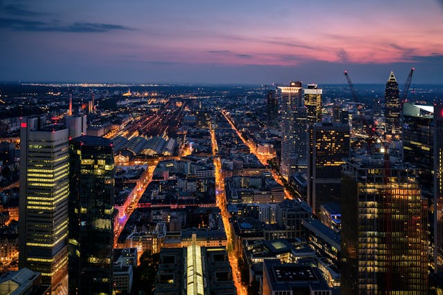 Luftaufnahme einer Stadtlandschaft in der Abenddämmerung mit beleuchteten Wolkenkratzern und Straßen. Der Horizont leuchtet in einer Mischung aus Orange- und Lilatönen, während die Lichter der Stadt darunter lebendige Muster erzeugen, die auf Frankfurts pulsierendes Nachtleben hindeuten, das durch die urbane Nacht pulsiert.