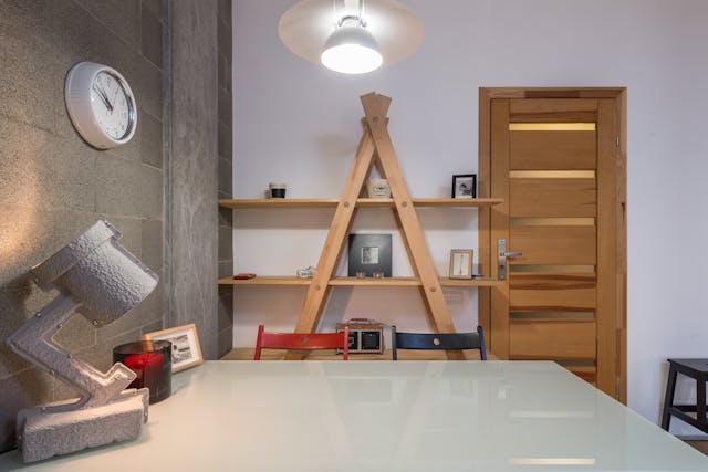 Ein modernes Bürointerieur mit einem weißen Tisch, einer einzigartigen Lampe und einer Wanduhr. Die Holztür verleiht dem umweltfreundlichen Zuhause Wärme, während Regale mit Dekoartikeln in Pyramidenform nachhaltige Wohnideen widerspiegeln. Der Raum verkörpert ein minimalistisches und stilvolles Design.