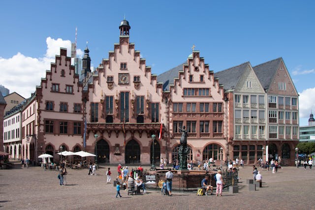 In Frankfurt am Main lädt ein belebter Platz zwischen historischen Gebäuden mit Treppengiebeln und verzierten Fassaden zum Verweilen und Flanieren ein. Ein Brunnen und eine Statue zieren das Zentrum unter einem teilweise bewölkten blauen Himmel und erinnern an Szenen aus einem malerischen YouTube-Video.