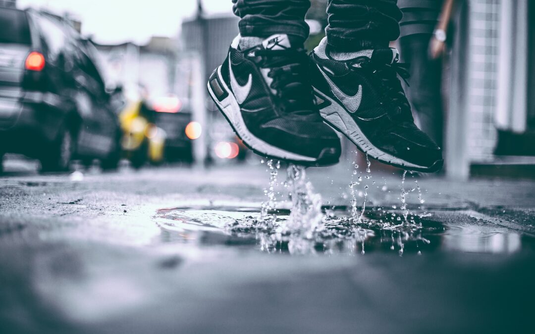 Eine Person in schwarzen Nike-Turnschuhen, die einen aktiven Lebensstil pflegt, springt über eine Pfütze auf einer verregneten Stadtstraße und spritzt dabei Wasser in die Luft. Die verschwommene Szene zeigt geparkte Autos und leuchtende Scheinwerfer im Hintergrund.