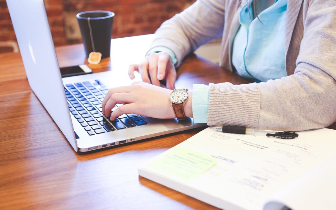 Eine Person in hellblauem Hemd und grauer Strickjacke tippt an einem Holztisch auf einem Laptop und plant wahrscheinlich eine Strategie für kommende Preisverhandlungen. Am Handgelenk der Person trägt sie eine Uhr. In der Nähe befinden sich ein offenes Notizbuch, ein Stift und eine schwarze Tasse mit Tee.